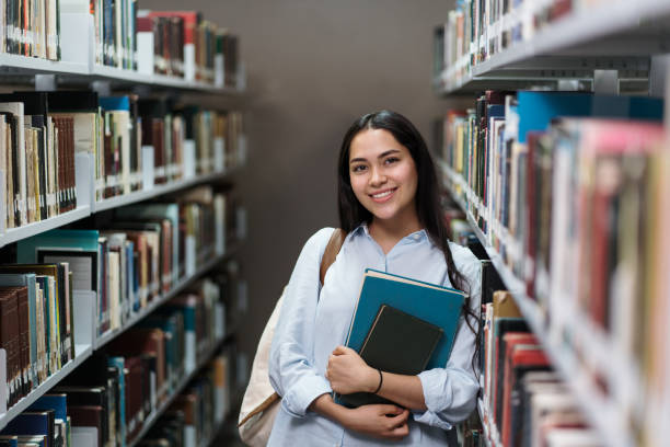 Happy female student leaning on bookshelves at library stock photo