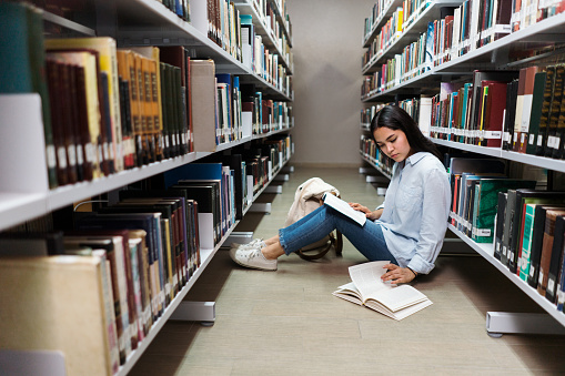 A latin female student sitting on the floor with book at the library.