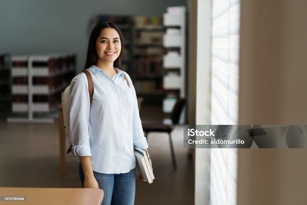 Female student standing with books in library and smiling at camera A latin female student standing with books by the window in a library and smiling at the camera. Student Stock Photo