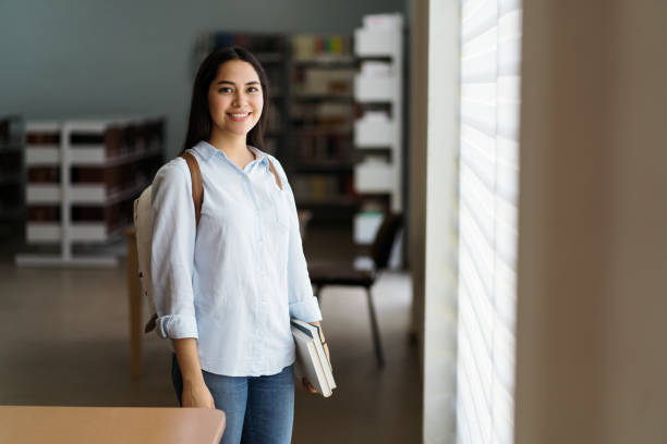Female student standing with books in library and smiling at camera stock photo