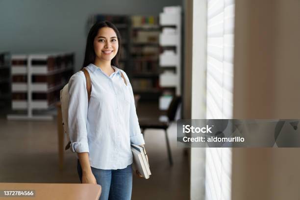 Female Student Standing With Books In Library And Smiling At Camera Stock Photo - Download Image Now