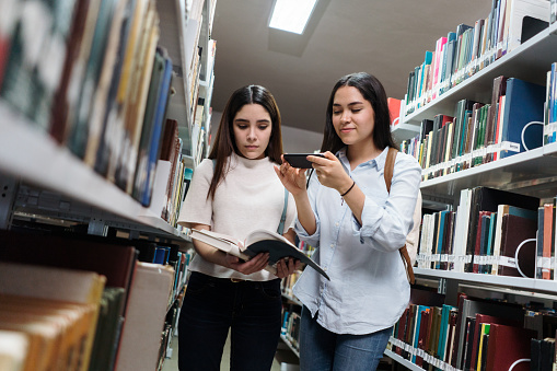 A female student standing and holding an open book for another student to take a photo with a mobile phone in a library.
