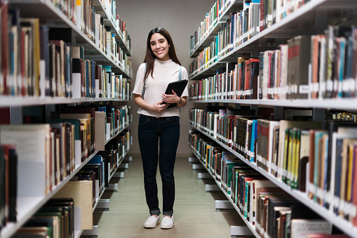 Friendly female student standing between bookshelves in library