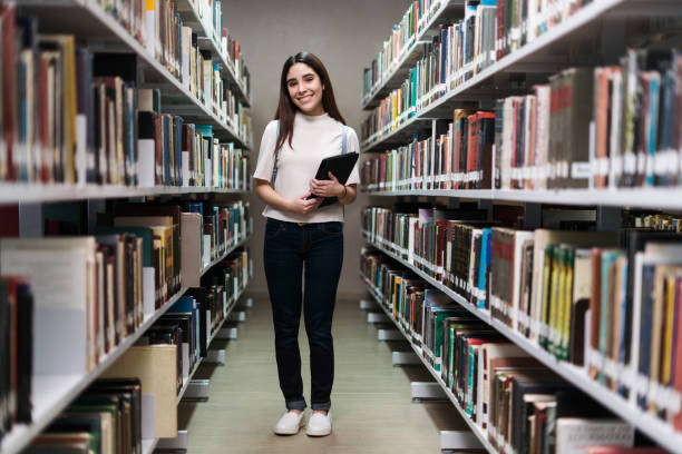 Friendly female student standing between bookshelves in library stock photo