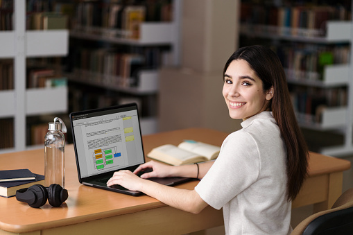 A happy latin female student typing on the computer at the library and smiling at the camera.