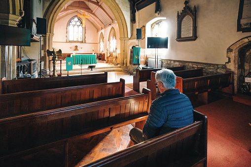 Color image depicting a senior caucasian man in his 60s in quiet prayer inside an old anglican church. The man has his hands clasped together in prayer. The man is alone and has his head bowed in solemn thought. Room for copy space.