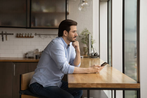 Peaceful businessman sit in office kitchen hold smartphone daydreaming