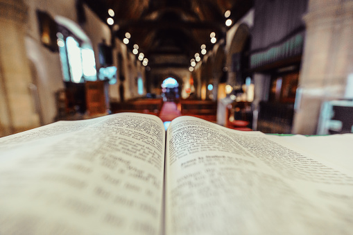 Color image depicting a close up wide angle view of the pages of an open bible on the altar of an Anglican church. Focus is on the bible in the foreground, while the background consists of the defocused interior of the church. Wooden pews recede into the distance and the lights illuminating the church appear as out of focus balls of light. The church has a warm, welcoming orange glow. Lots of room for copy space.