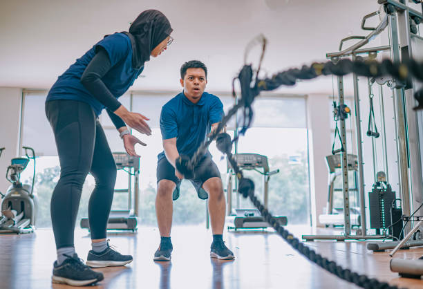 Asian Malay female personal trainer encouraging Asian Malay man practicing challenging battle rope in gym Asian Malay female personal trainer encouraging Asian Malay man practicing challenging battle rope in gym fitness instructor stock pictures, royalty-free photos & images