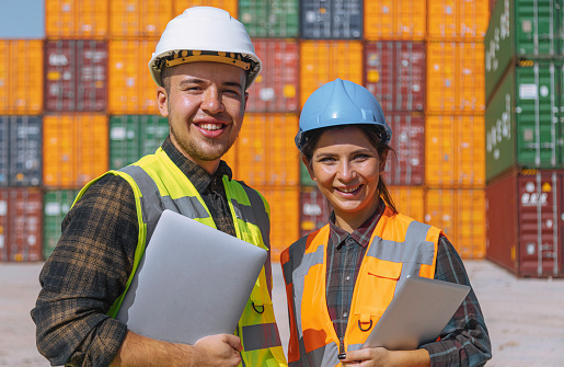 Portrait of happy young business team of male transport engineer foreman and businesswoman wearing protective white and blue crash safety helmets and worker wests holding laptop computer and digital tablet notebook smiling in front of colorful cargo container stacks in shipping port on large commercial dock. Transportation and logistics concept working lifestyle