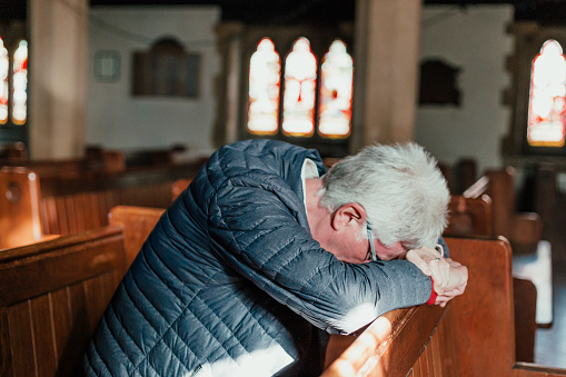 Color image depicting a senior caucasian man in his 60s in quiet prayer inside an old Anglican church. The man has his hands clasped together in prayer. The man is alone and has his head bowed in solemn thought. Room for copy space.