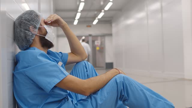 Exhausted doctor in medical mask and cap having headache sitting on floor in hospital corridor