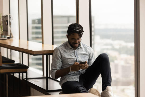 istock Relaxed African ethnicity man using smartphone, sitting on windowsill. 1372801420