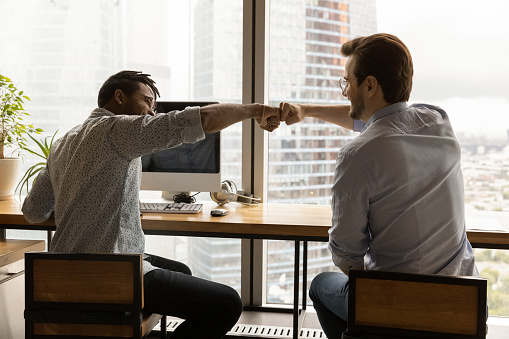 Happy two male colleagues bumping fists, greeting each other or showing team union. Joyful young business partners celebrating shared success, sitting at table in modern workplace.