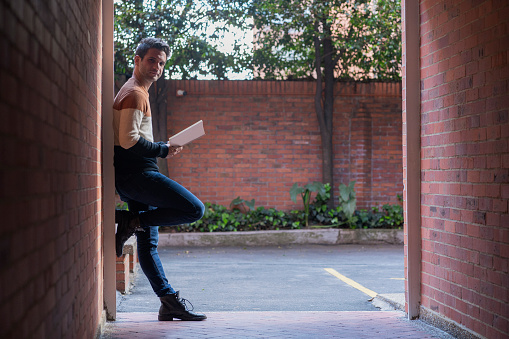 A 29-year-old Latino man from Colombia, Bogotá, casually dressed, stands outside his building reading a book in the open air while looking at the camera that portrays him