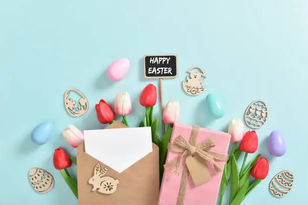 Photo of A bouquet of tulips with a postcard, a gift and wooden Easter eggs with a plaque on a light blue background.