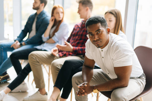 istock Tired African-American young man job candidates looking at camera while waiting interview with hr, sitting in queue line row with bored multiethnic candidates. 1372809408