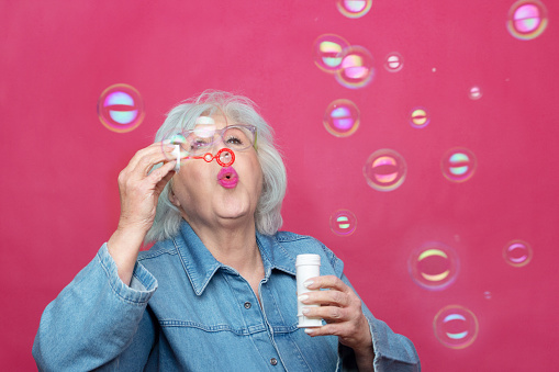 grey haired senior woman blowing soap bubbles on a plain background