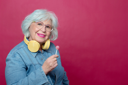 fashionable older woman with headphones around her neck holding a lollipop looking at camera on pink background