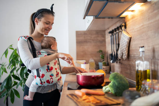 Multi-task mother cooking and taking care of newborn son. Multi-tasking mother in the kitchen cooking and taking care of her newborn son. baby carrier stock pictures, royalty-free photos & images