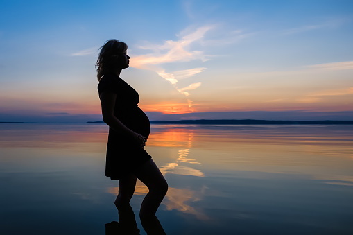 Full length silhouette unrecognized female expecting a baby, who stands in the water of the lake and touch her belly against evening sky background at sunset. Pregnancy, copy space