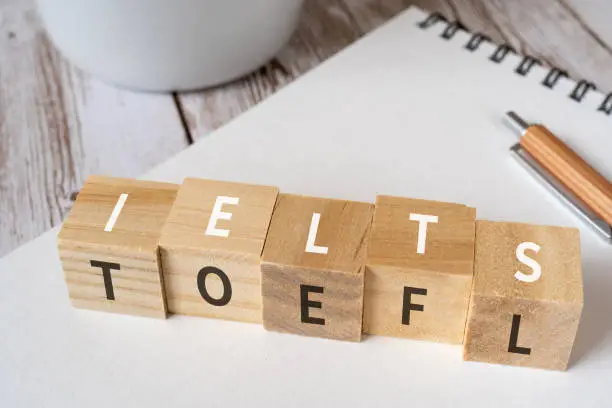 Wooden blocks with "IELTS" and "TOEFL" text of concept, a pen, a notebook, and a cup.