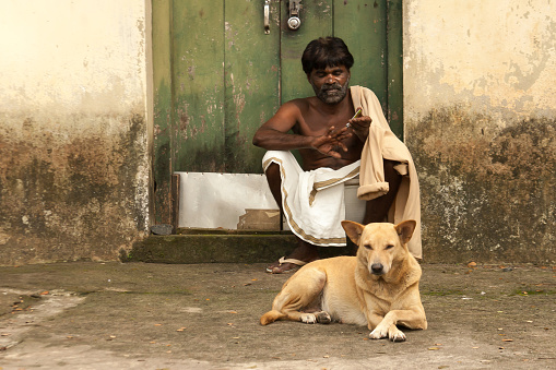 Kochi (Kerala), India: A man sitting on a stoop at a bathhouse in downtown Kochi trimming his nails, with a dog at his feet.