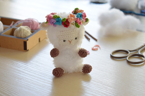 Making fanny sheep with wreath of flowers. Crochet toy ewe for child. On table threads, needles, hook, cotton yarn. Handmade crafts. DIY concept. Small business. Income from hobby