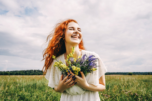 Young beautiful curly ginger woman holds flowers in her hands and laugh. Field and sky on background.