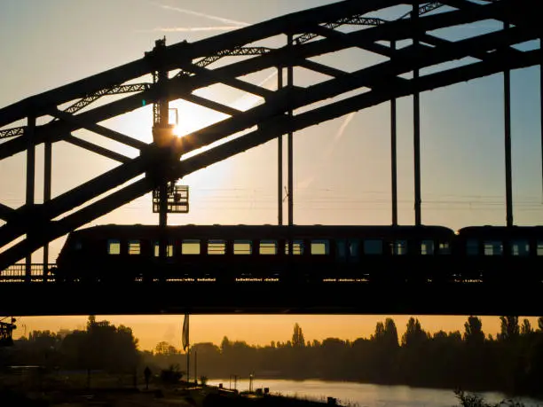 Silhouette of a passenger train crossing a metal arch bridge in the backlight of the rising sun