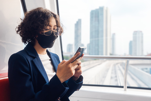 Curly girl Businesswoman wearing a mask uses her phone while taking a sky train, people lifestyle with technology devices and telecommunication concept