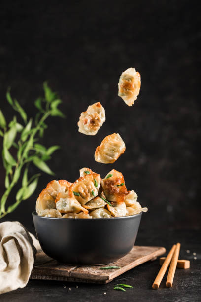 Fried dumplings with pork and herbs in a black round bowl and flying in the air Fried dumplings with pork and herbs in a black round bowl and flying in the air on a dark background with chopsticks. Korean food. Side view with copy-paste for text. Vertical orientation. chinese dumpling stock pictures, royalty-free photos & images