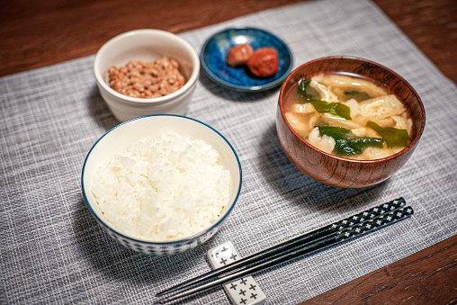 A traditional Japanese lunch set teishoku with a bowl of white rice, miso soup, fermented beans natto, and pickled plums umeboshi served with a pair of black chopsticks on a striped grey table cloth