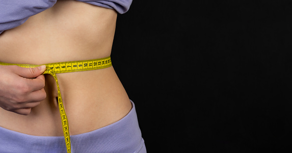A slender girl measures her waist with a measuring tape. Healthy body, weight loss concept. Slim waist, small belly in lilac sports pants and a T-shirt. Black background.