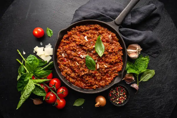 classic italian bolognese sauce stewed in a pan with ingredients on black tile background, top view