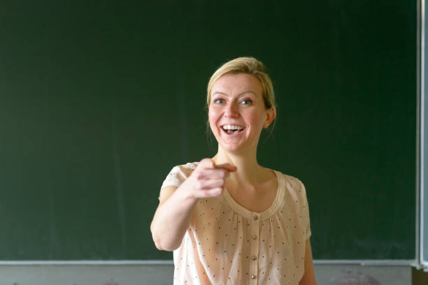 Schoolteacher standing in front of a blackboard in the classroom pointing with her finger saying You