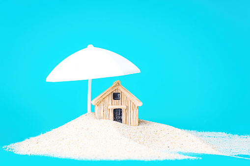 Miniature toy house standing in a sand pile under a white parasol isolated on blue. Creative home erosion protection concept.
