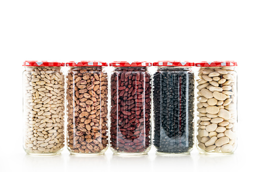 Assorted beans legumes in glass jar in a row on white background as: kidney beans, white beans, black beans, pinto beans, red beans