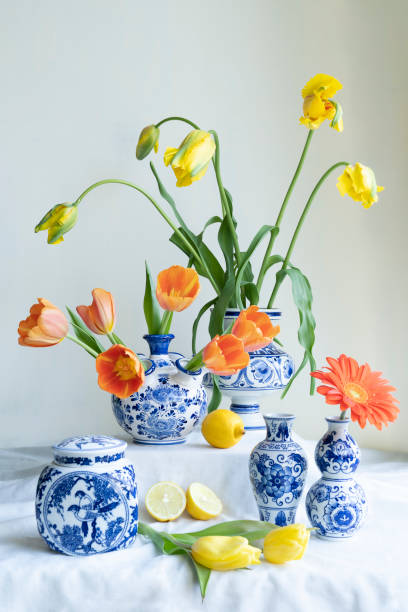 un exuberante bodegón de luz natural con un viejo jarrón holandés con tapa azul de delft con tulipanes amarillos y naranjas, una gerbera y limones sobre lino blanco sobre un fondo blanco. - delftware fotografías e imágenes de stock