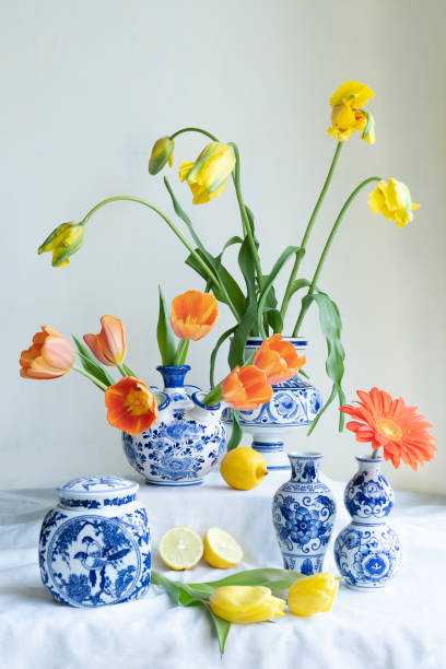 uma luz natural exuberante ainda cheia com o velho vaso azul delft holandês com tulipas amarelas e laranjas uma gerbera e limões em linho branco contra um fundo branco. - delftware - fotografias e filmes do acervo