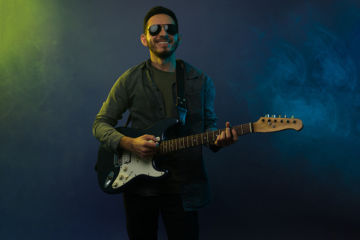 Excited hispanic man smiling while playing the electric guitar against a background with white smoke