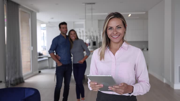 Female Real Estate Agent looking at tablet and then facing camera while an adult couple stand at background all smiling