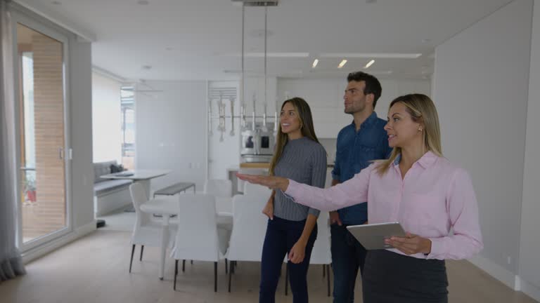 Cheerful female Real Estate Agent holding a tablet and showing a beautiful modern property to happy adult couple