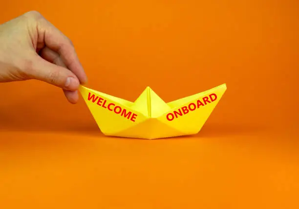 Photo of Welcome onboard and support symbol. Concept words Welcome onboard on yellow paper boat on a beautiful orange table orange background copy space. Businessman hand. Business and Welcome onboard concept.