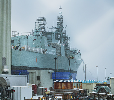 The superstructure and hull of HMAS Adelaide, a Canberra Class amphibious assault ship of the Royal Australian Navy docked at Garden Island, Sydney Harbour.  In the background are two of the dockside cranes. The safety netting around the deck casts a shadow over the hull.  This image was taken on a hot and sunny afternoon on 6 January 2024.