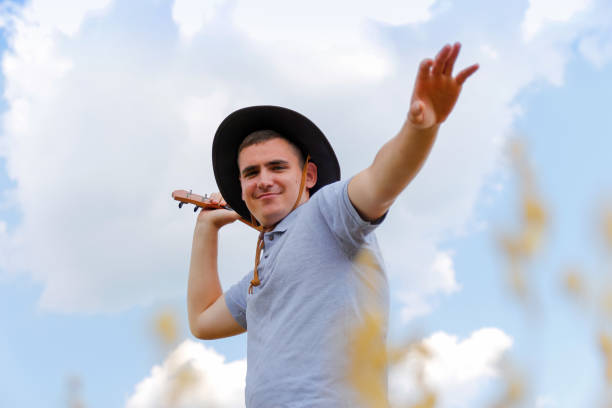 músico de emoción. joven hermoso hombre caucásico tocando el ukelele afuera. el guapo chico hipster toca la guitarra de ukelele en el fondo de la naturaleza. hombre con sombrero de vaquero sonríe y toca la guitarra - cowboy blue meadow horizontal fotografías e imágenes de stock