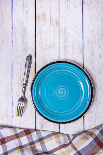 istock Top view background with empty blue ceramic plate, antique silver cutlery on vintage weathered white wooden boards. Copyspace. Brown checkered kitchen towel. Vertical photo. 1373028901
