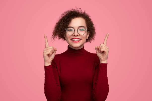Cheerful young woman pointing up and smiling at camera Excited young female model with curly hair in turtleneck and eyeglasses, smiling and looking at camera while pointing up with index fingers against pink background high collar stock pictures, royalty-free photos & images