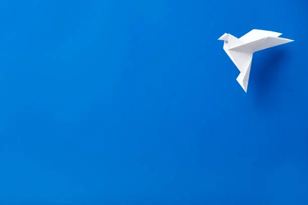 Photo of White origami paper dove flying against a blue background.