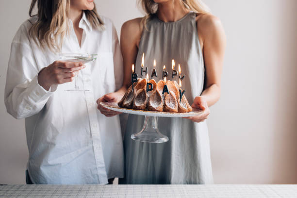 Happy Birthday! Two Unrecognizable Female Friends Celebrating, Holding a Gugelhupf with Candles and a Coupe Glass Two anonymous female friends celebrating a birthday, holding a bundt cake with candles and a coupe glass. woman birthday cake stock pictures, royalty-free photos & images